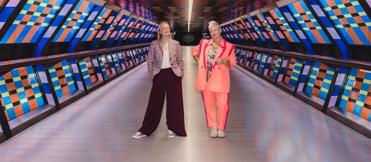Two women standing in a brightly lit tunnel with colorful geometric patterned walls and ceiling. One woman wears a light pink jacket and dark purple wide-leg pants, while the other wears a bright coral-pink pantsuit.